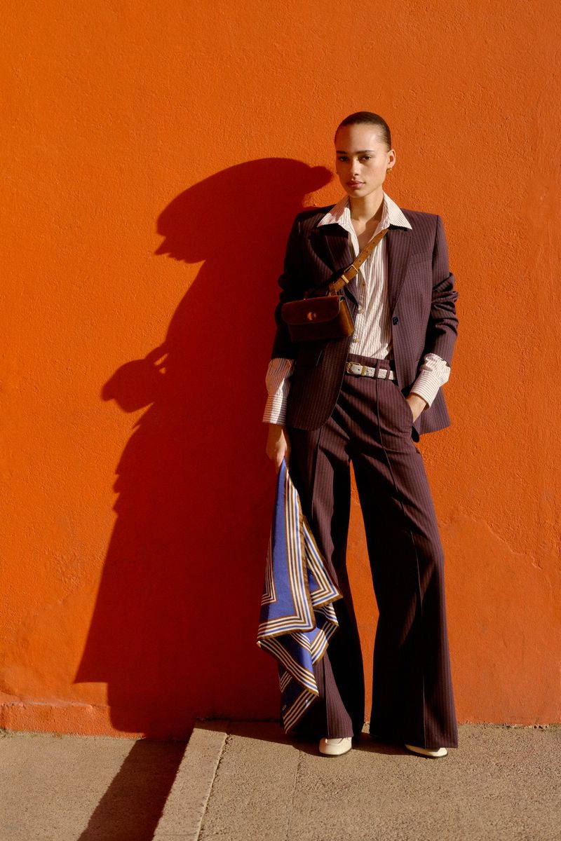 A stylish young woman in a pinstripe suit poses against a vibrant orange wall, accessorized with a brown purse and holding a blue scarf.