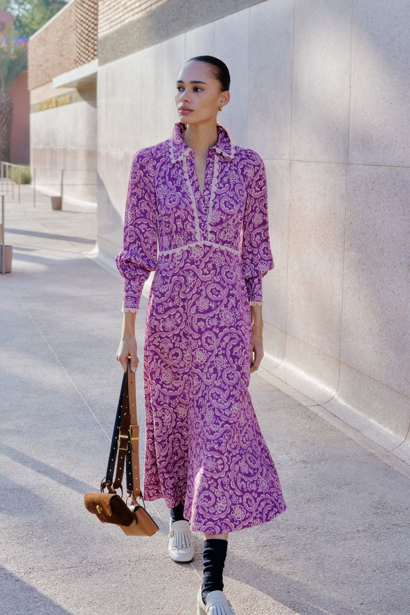 Adult female in a purple floral dress with a shoulder bag poses on a sidewalk next to a wall and planters with sunlight.
