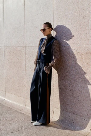 Model leaning against a textured stone wall wearing a navy vest, patterned blouse, wide-leg trousers, and sunglasses.
