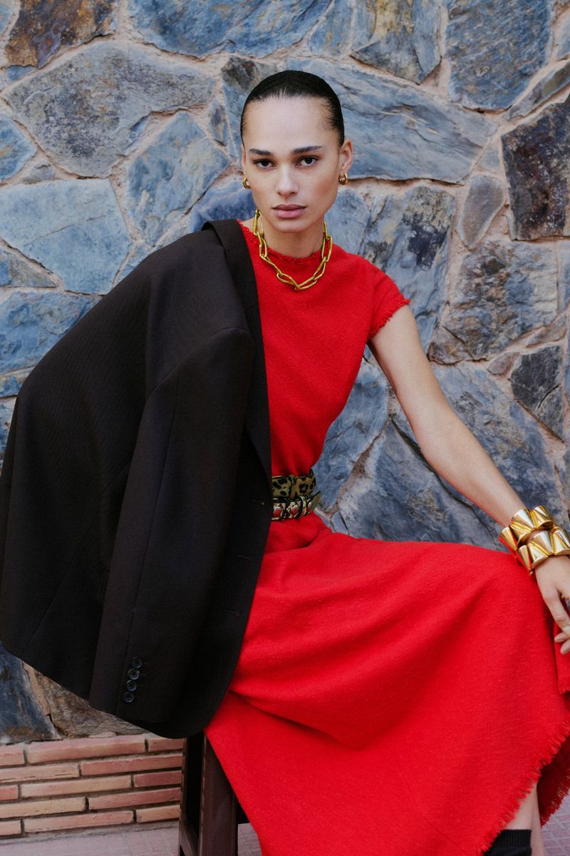 Model in a red dress with a dark jacket draped over her shoulder posing against a textured stone wall