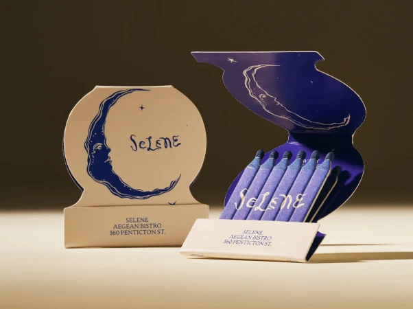 Two matchboxes are displayed, one closed and one open with blue-tipped matches, both featuring the logo for Selene Aegean Bistro.