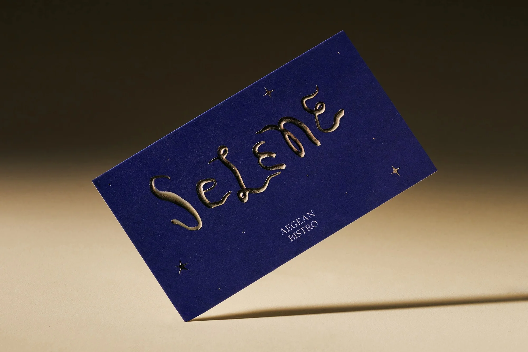 Close-up of a navy business card with gold embossed lettering that reads 'Selene Aegean Bistro' on a neutral background with a light source.