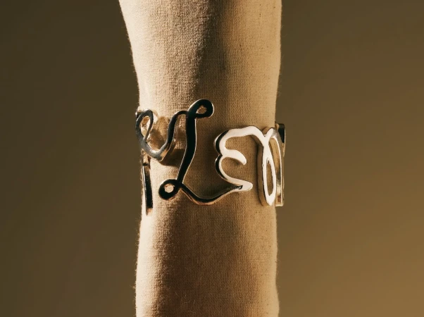 Close-up of a silver metal cursive word-design bracelet around a beige napkin roll, soft lighting, neutral studio background.