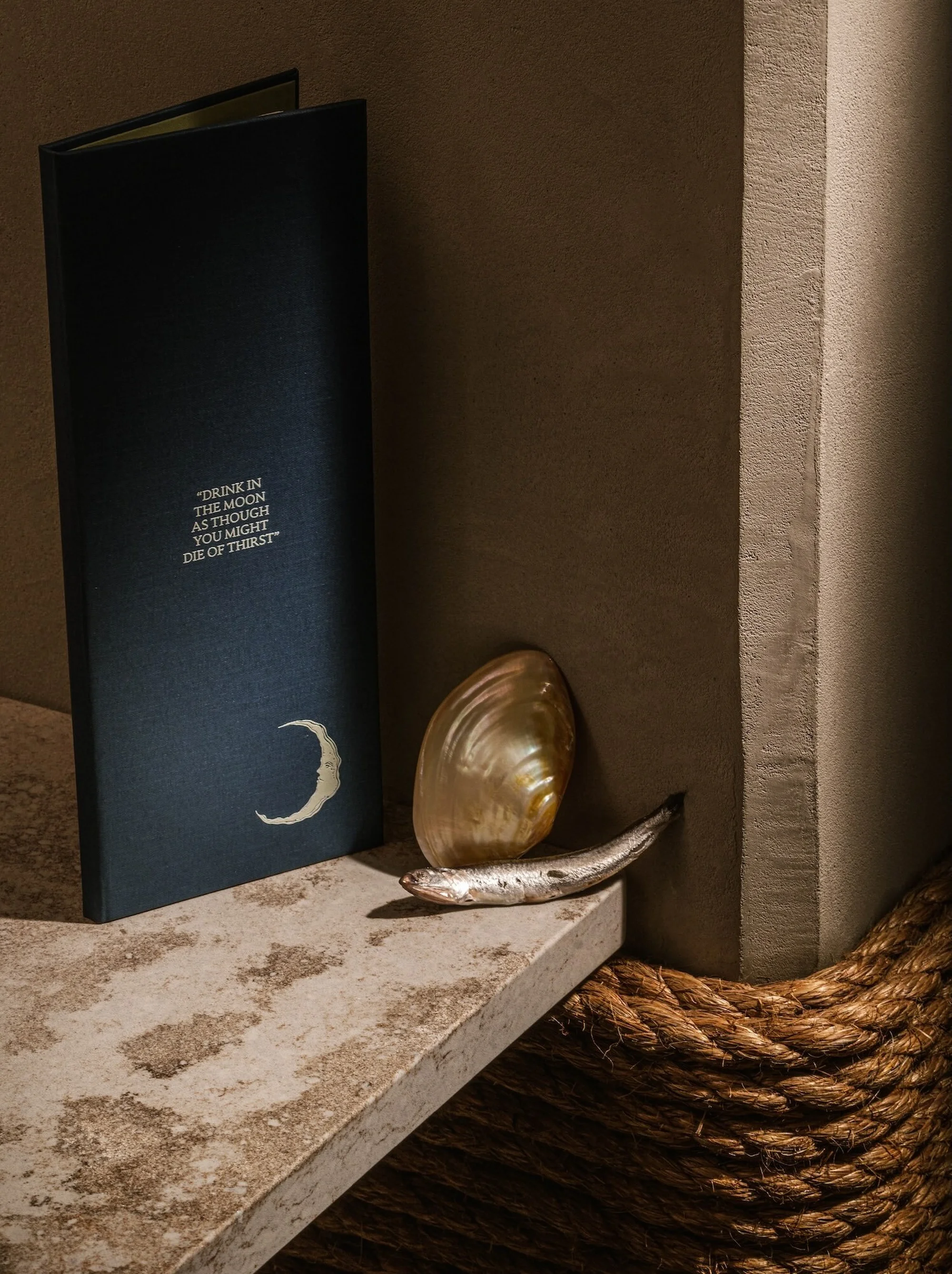 Navy menu with a crescent moon design sits next to a glistening shell and a small fish on a stone ledge with a rope accent.