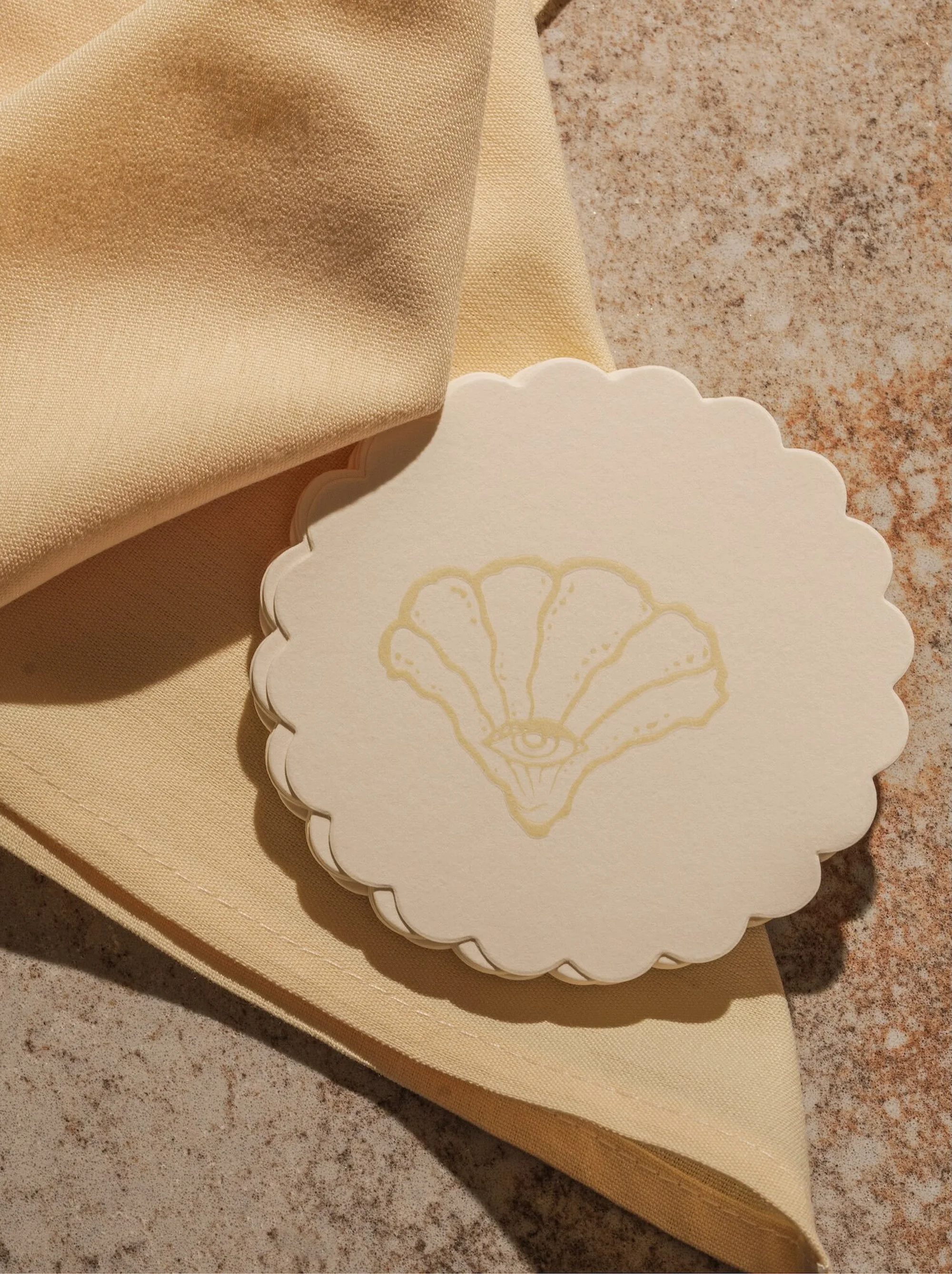 Beige table setting featuring a scalloped-edged coaster printed with an eye-centered seashell design beside a soft, folded cloth napkin.