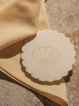 Beige table setting featuring a scalloped-edged coaster printed with an eye-centered seashell design beside a soft, folded cloth napkin.
