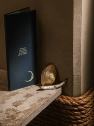 Dark blue menu with silver foil moon and quote resting on a textured stone ledge beside a shell and a small silver object.