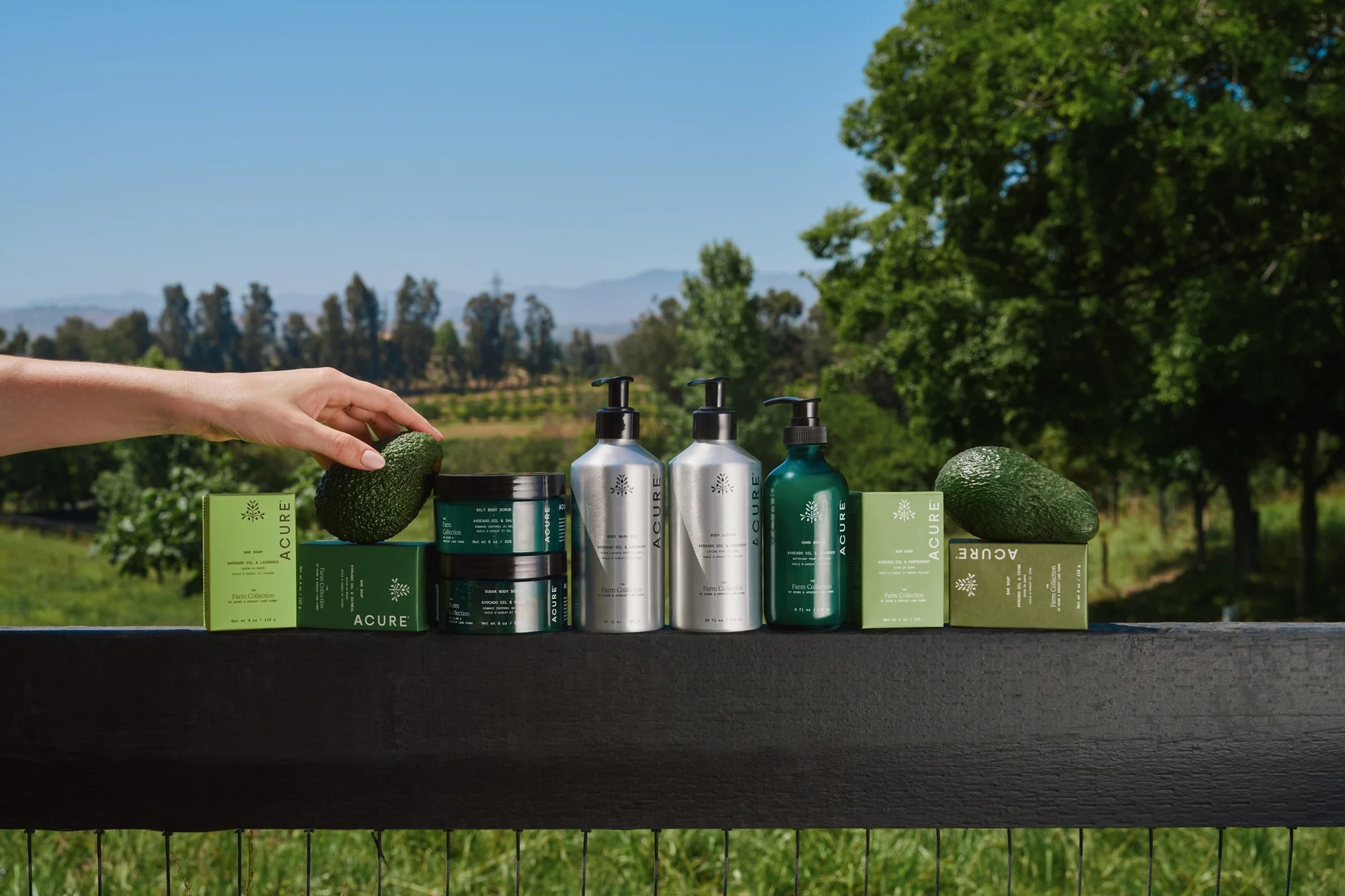 Cosmetic products from Acure sit on a black ledge with an avocado and a hand touching it against a clear blue sky backdrop.