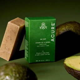 Acure green bar soap box flanked by a soap bar and fresh avocados on green background