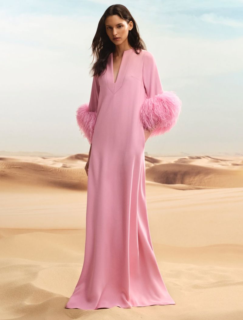 A woman in a flowing pink gown with fur cuffs stands in a desert landscape under a bright sky in a fashion editorial.