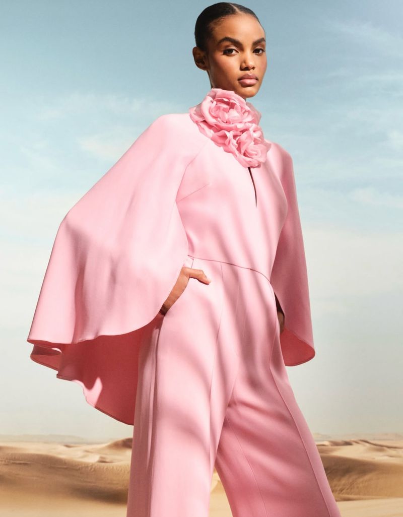 Fashion model in pink jumpsuit with large flower collar, poses in front of sand dunes under a bright sky in a fashion editorial.