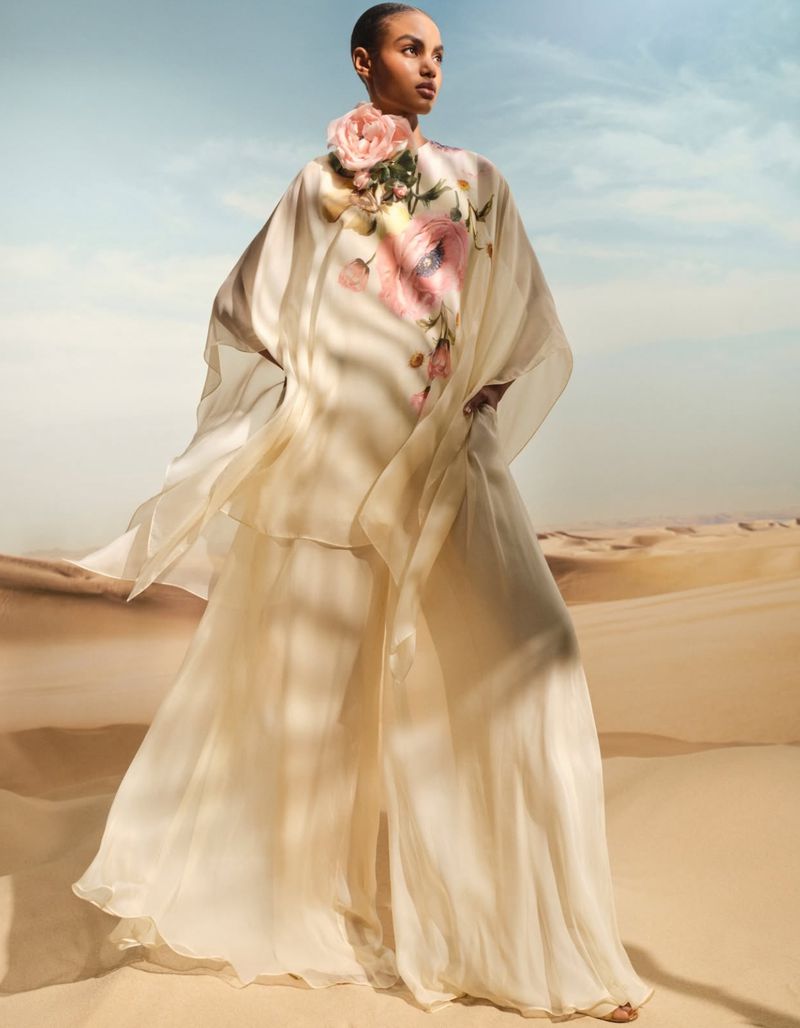 A woman models a sheer cream-colored outfit with a flower detail, posing in a desert with a blue sky, conveying a soft, elegant fashion style.