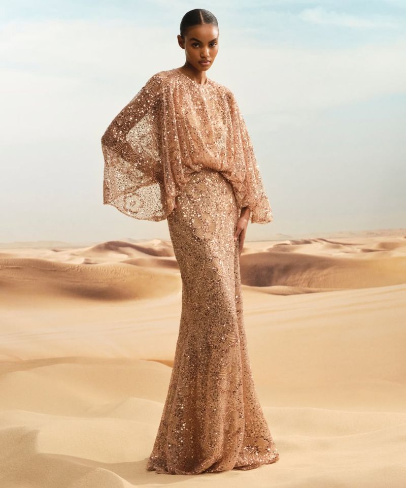 Elegant woman in a shimmering gold gown with flowing cape poses in a sunlit desert. Fashion photography with sandy dunes backdrop.