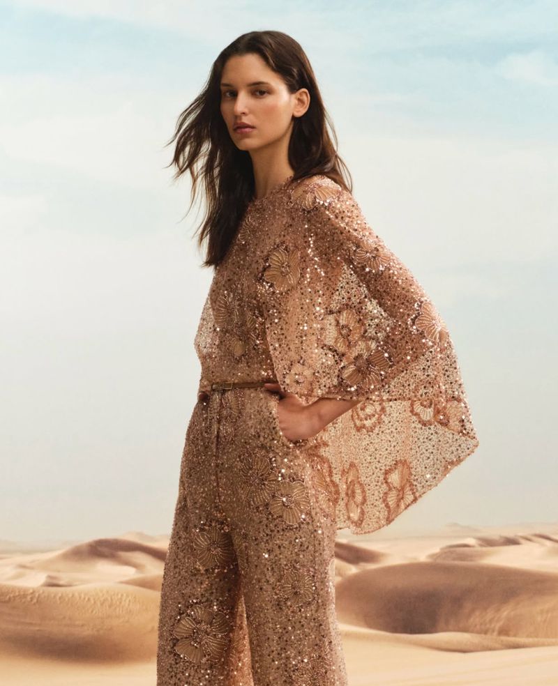 Woman in sequined top and pants posing in a desert setting. Fashion shot with neutral colors, clear sky, and sand dunes. Beauty and style.