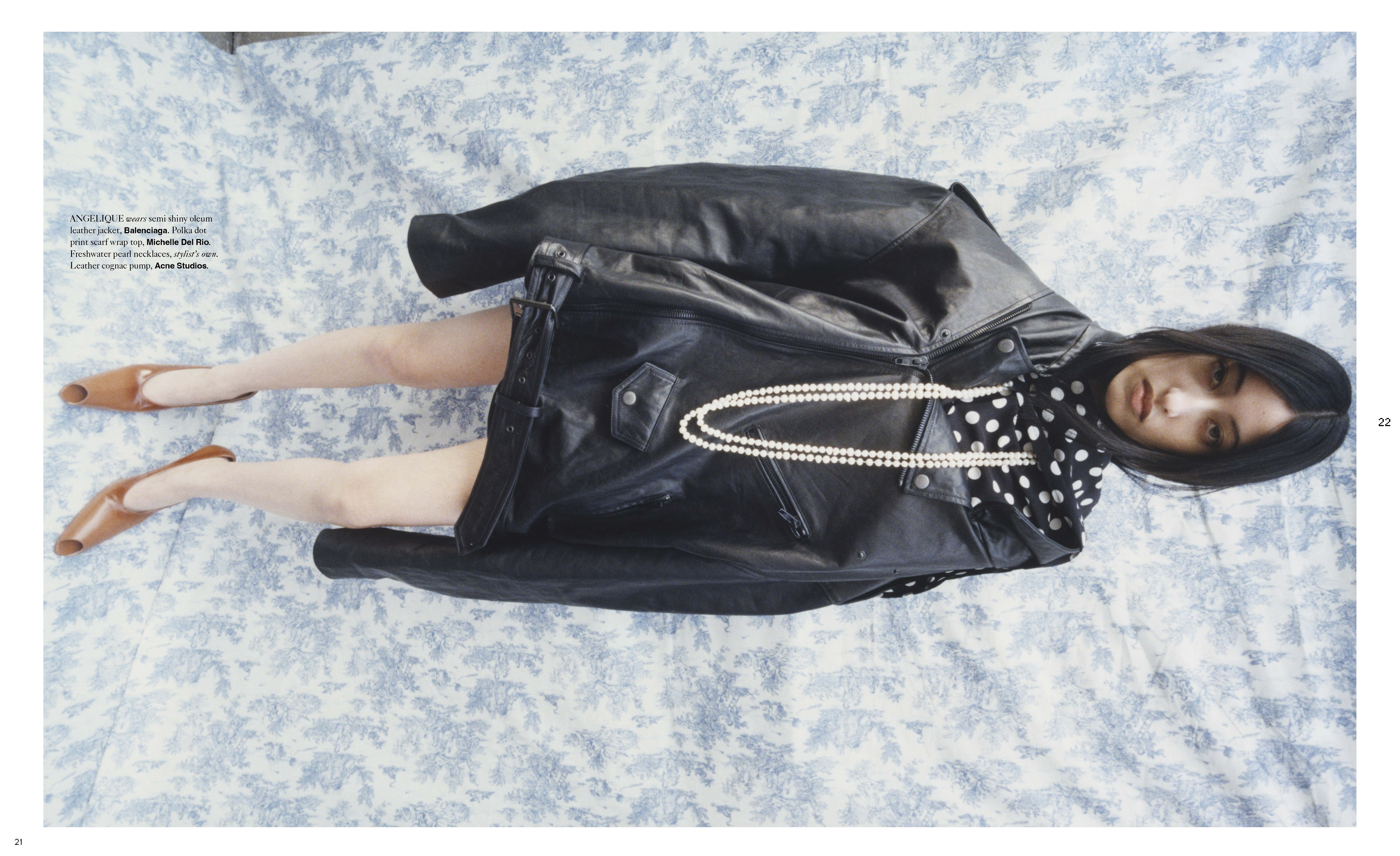 Fashion editorial shot with a brunette woman in a leather jacket and polka dot scarf lying on a floral-patterned surface, wearing heels and a pearl necklace.
