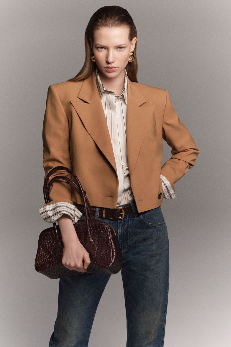 A woman in a tan blazer, striped shirt, and blue jeans holds a dark brown purse in a studio setting against a grey backdrop.