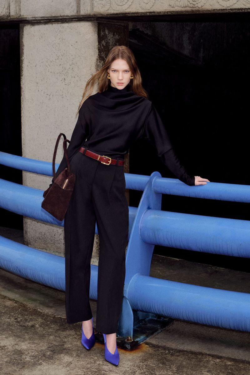 A single female model posing in fashionable black top and pants with purple heels, next to a blue painted railing. She is carrying a brown bag.