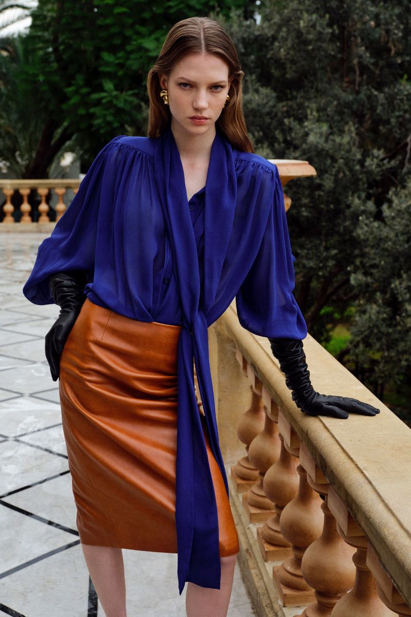 Stylish woman in a blue blouse and brown leather skirt posing outdoors with trees in the background, exuding fashion and elegance.