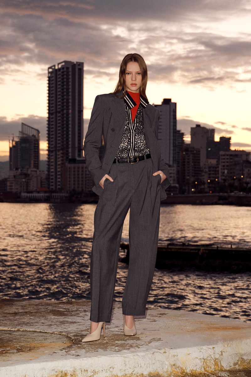 Fashion shot of a woman in a grey suit with a red top, standing on a concrete surface with water and a city skyline behind her.