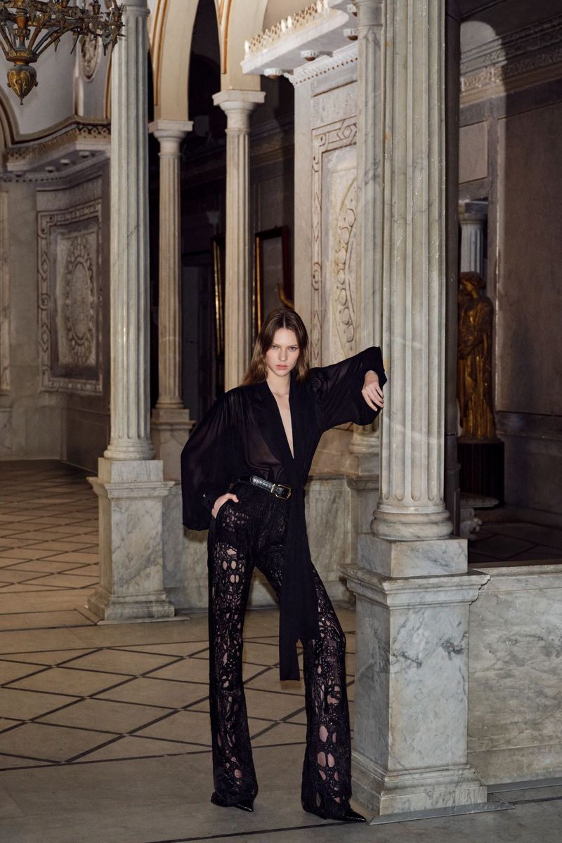 Fashion shot of a woman in a black top and lace pants, leaning against a marble column inside a vintage-style building with high ceilings.