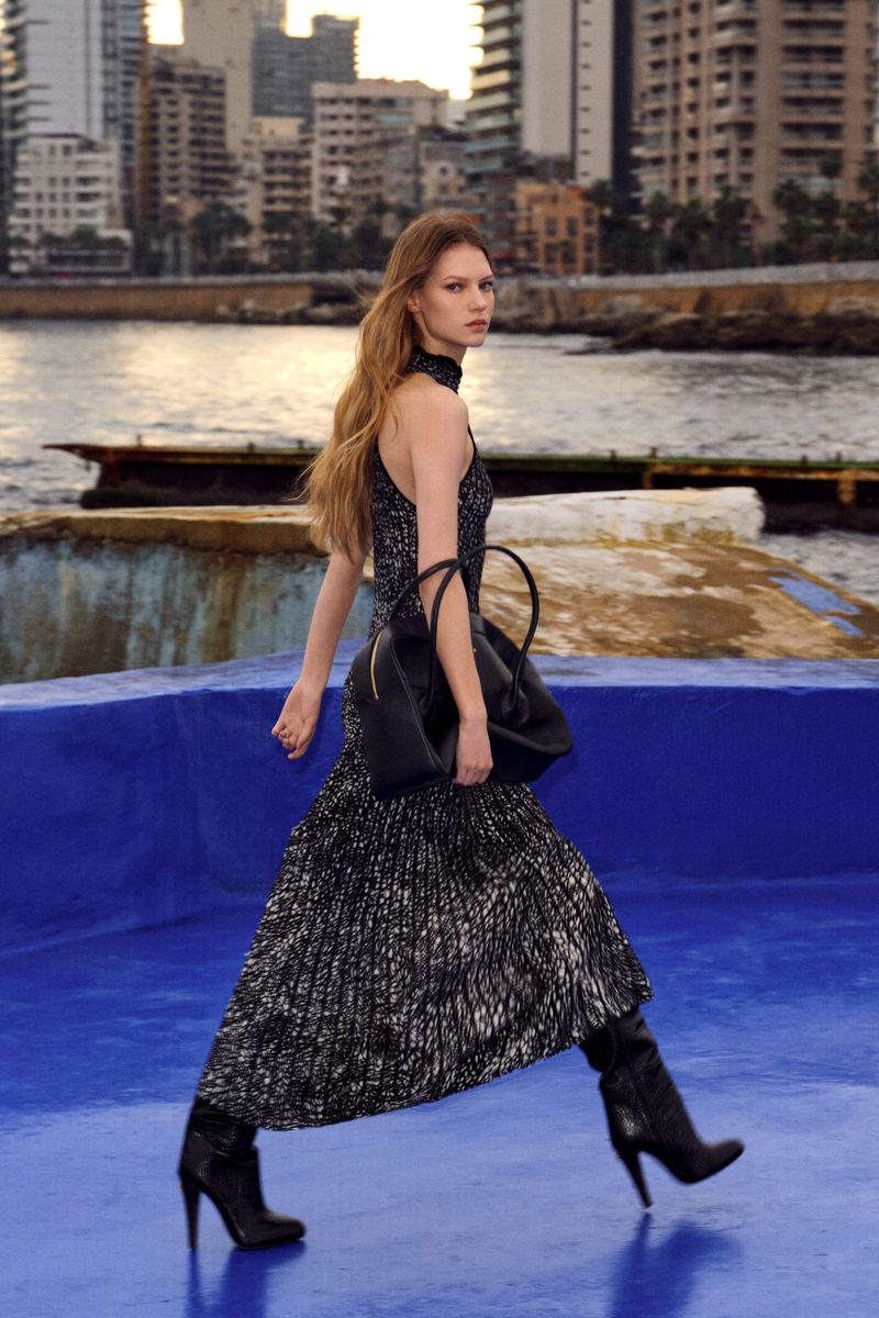 Fashion model walks on a blue surface with a city skyline in the background, wearing a dress, handbag and high heel boots.