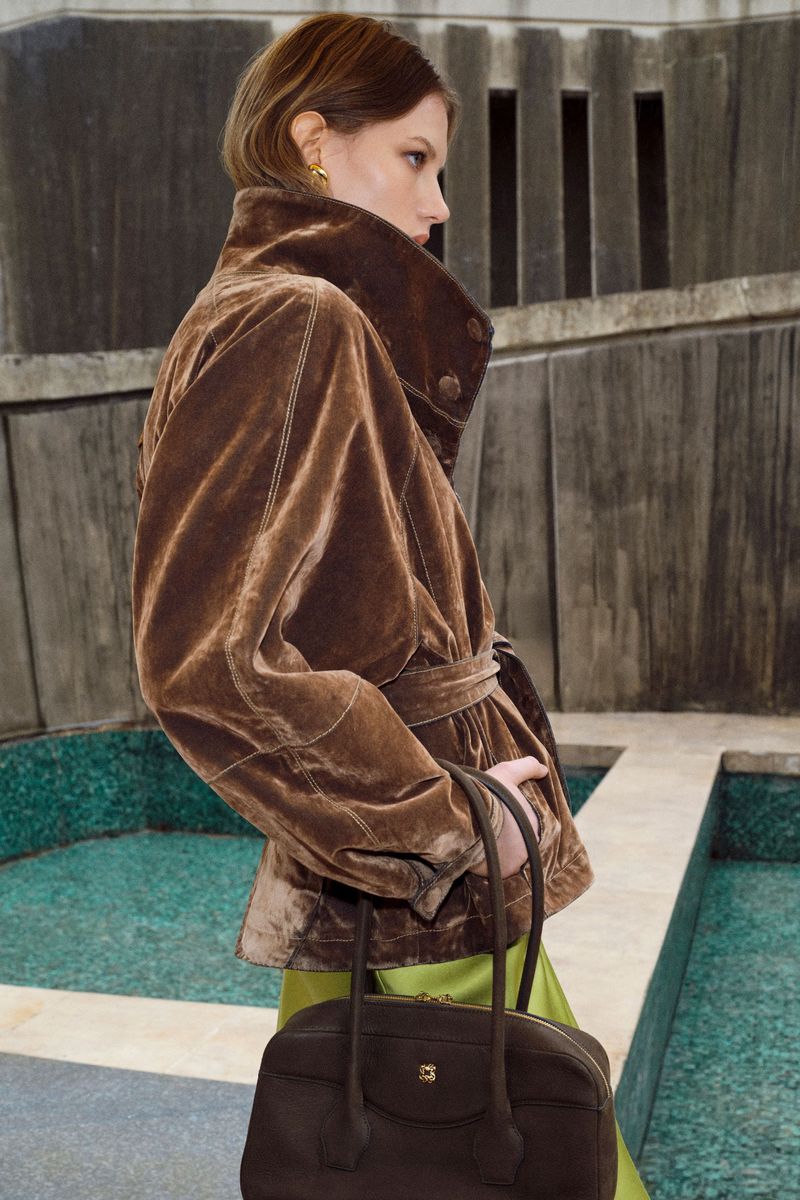 A model poses in a brown velvet jacket and green skirt, carrying a brown handbag in an indoor setting with patterned walls.