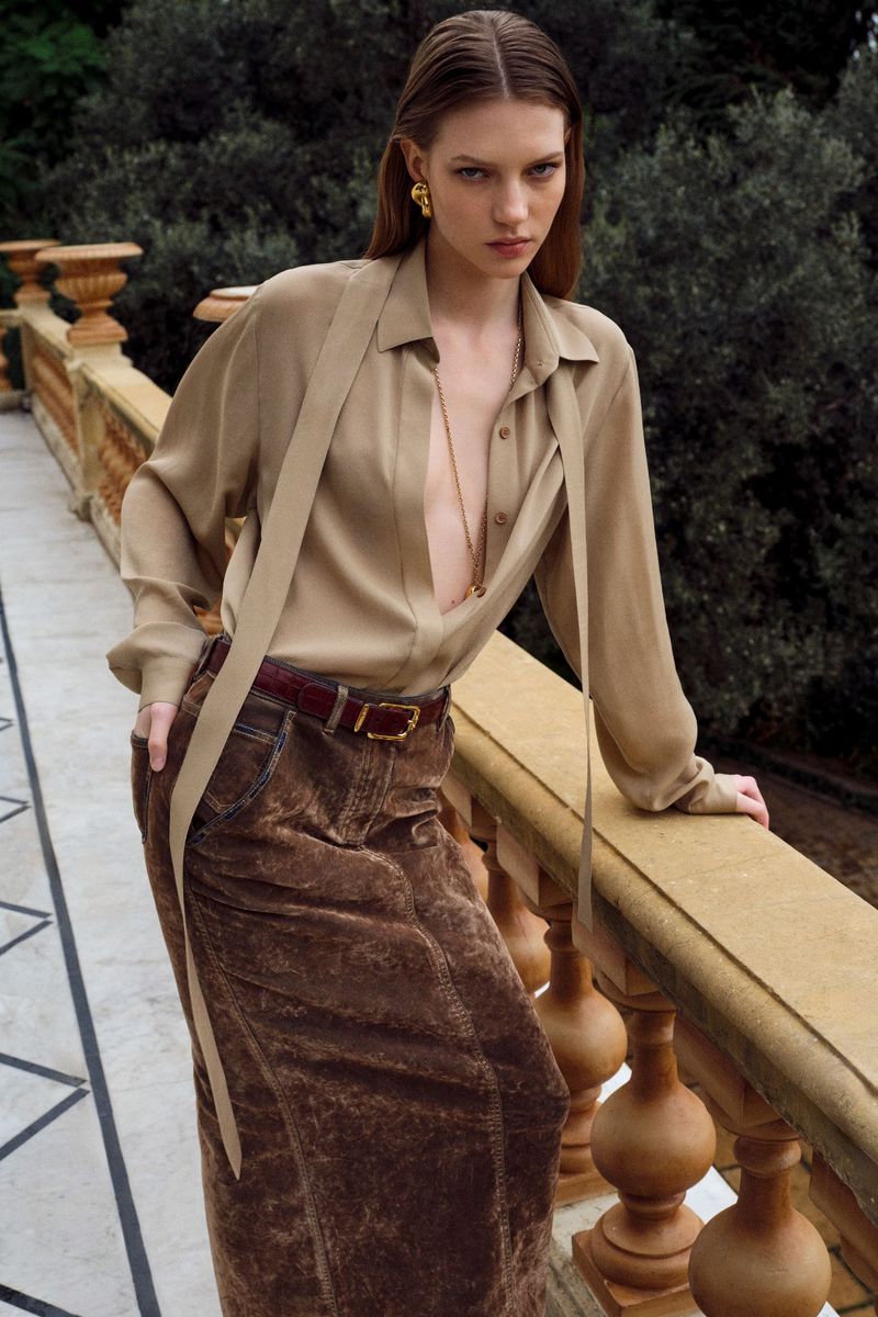 A poised adult female model in a tan blouse and brown skirt leans against a stone railing with greenery in the background.