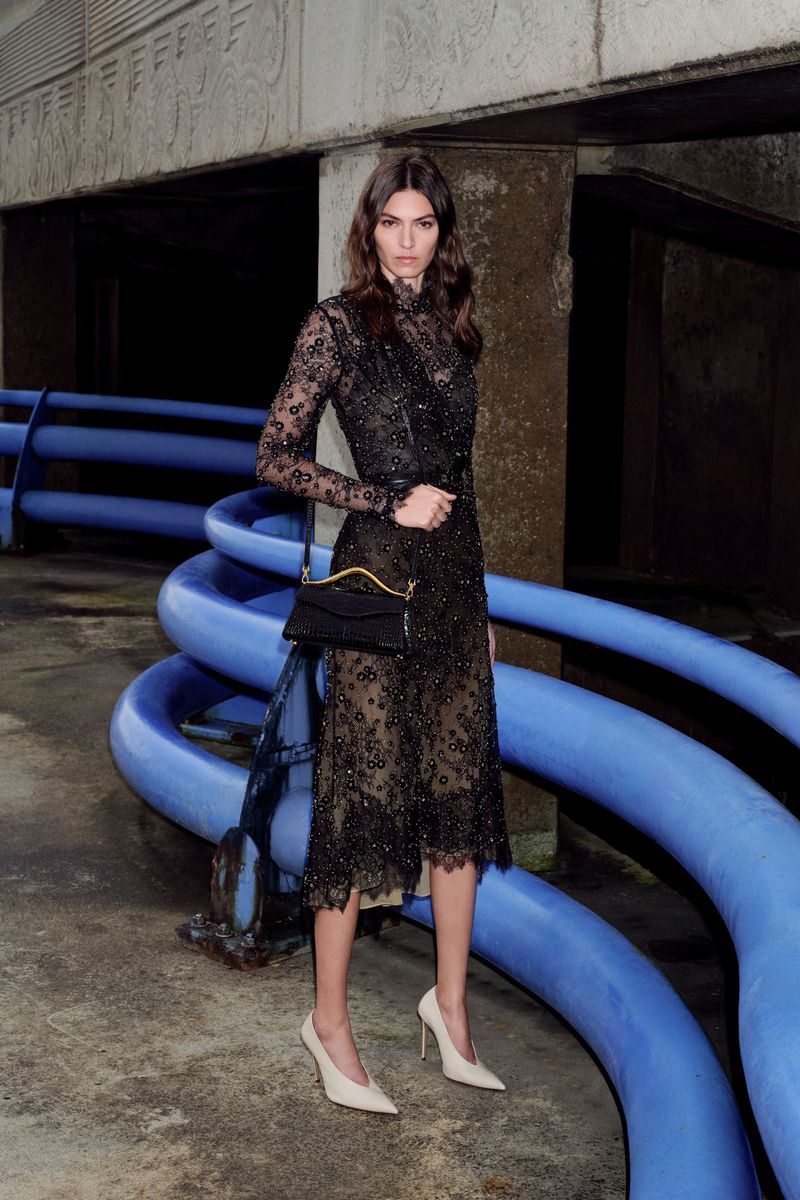 Adult female wearing a black lace dress and cream pumps in a concrete urban environment with blue pipes as a backdrop.