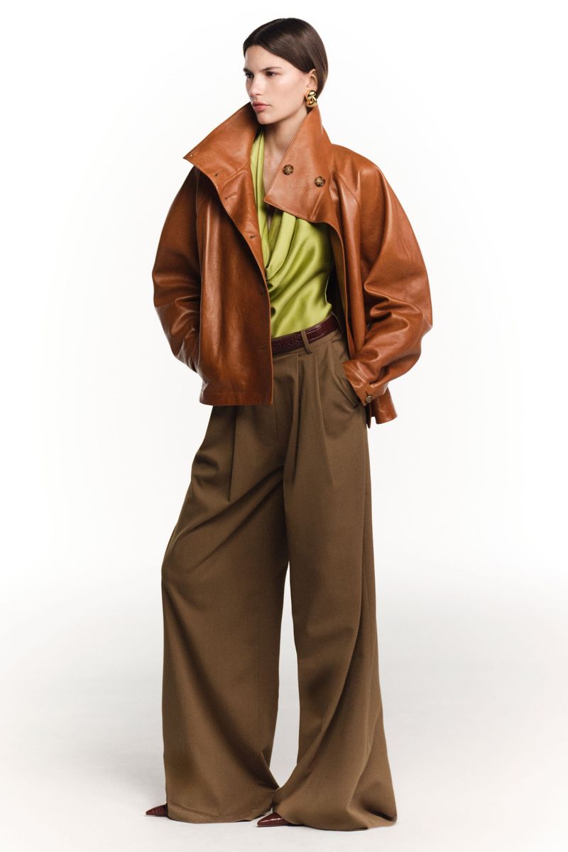 A model presents a fashionable ensemble featuring a brown leather jacket, a light green top, and brown wide-leg pants against a white backdrop.