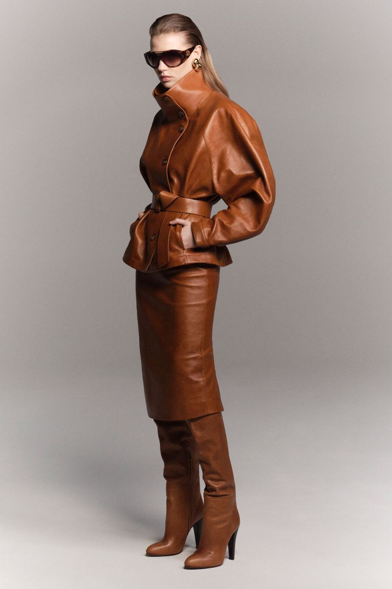 A woman wearing sunglasses and a brown leather jacket, skirt, and boots is posing in a studio. Fashion photography.