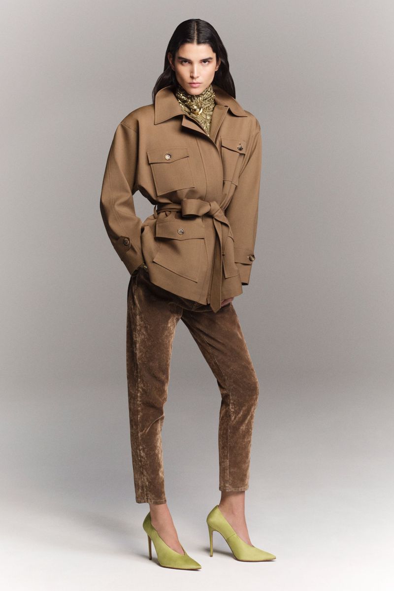 An adult woman in a brown jacket with a waist tie, brown trousers, and high heels stands in a photo studio with neutral background.