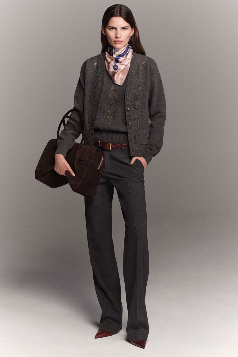 A woman models a neutral-toned outfit with a cardigan, sweater, wide-leg pants, and a brown handbag against a grey studio backdrop.