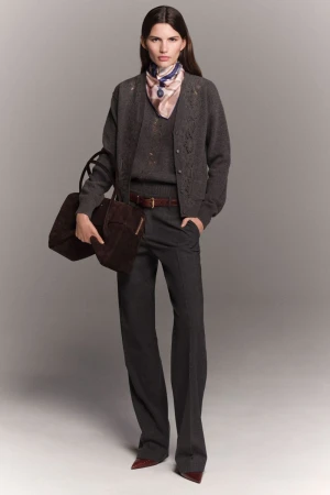 A woman models a neutral-toned outfit with a cardigan, sweater, wide-leg pants, and a brown handbag against a grey studio backdrop.