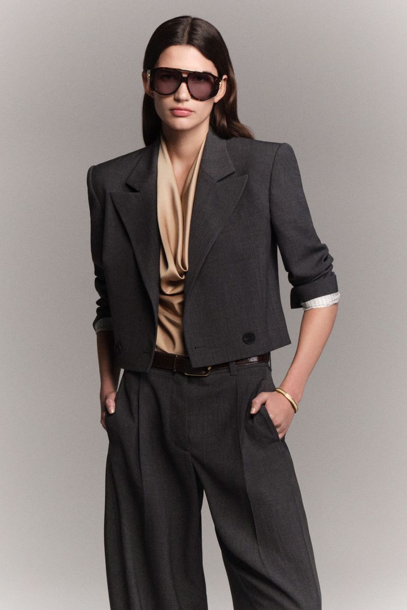 Stylish woman in a cropped blazer, blouse, and wide-leg trousers, complemented by sunglasses and a bracelet, set against a neutral backdrop.