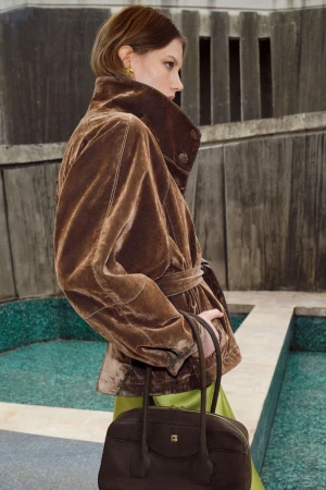 Model wearing a brown velvet belted jacket holding a dark brown handbag against a textured concrete and pool background.