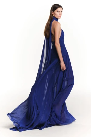Model in a flowing royal blue halter gown with a trailing cape against a white seamless background for Elie Saab Pre-Fall 2026.