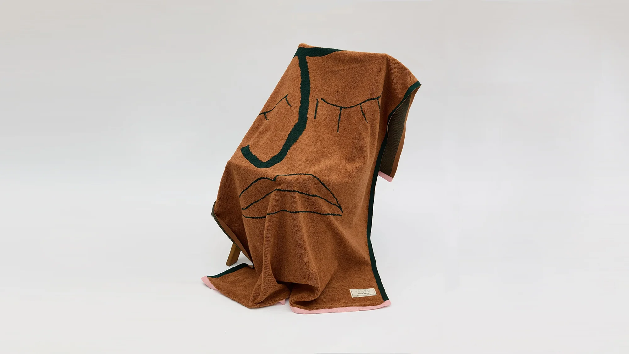 Brown knit blanket with a minimalist face design draped over a wooden chair against a plain white background in a studio setting.