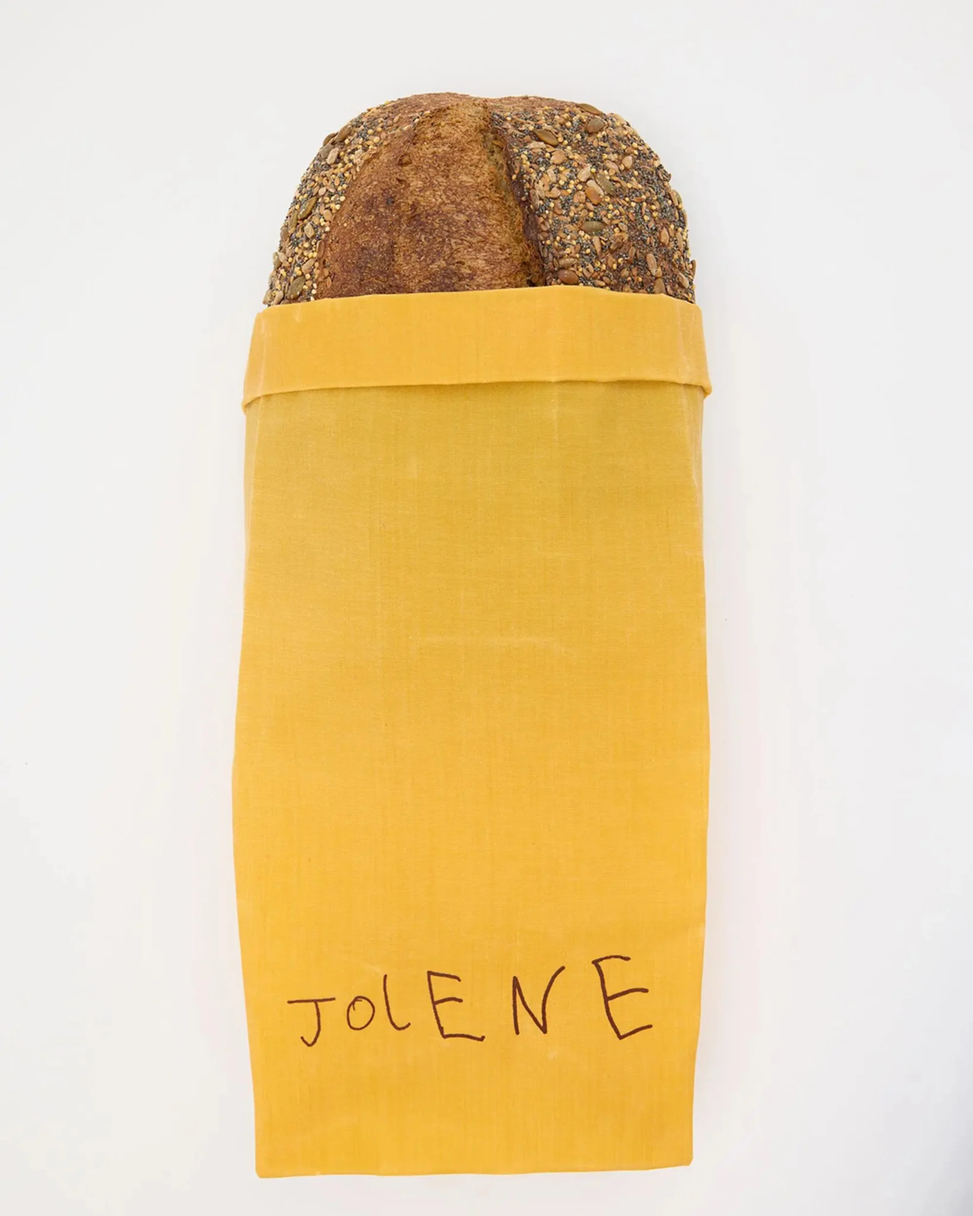 A loaf of bread in a yellow fabric bag with "Jolene" written on it, centered against a plain white background, studio lit.