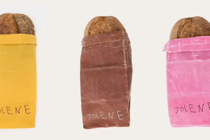 Three loaves of bread in pink, yellow, and brown reusable waxed canvas bags, standing side by side on a white surface.