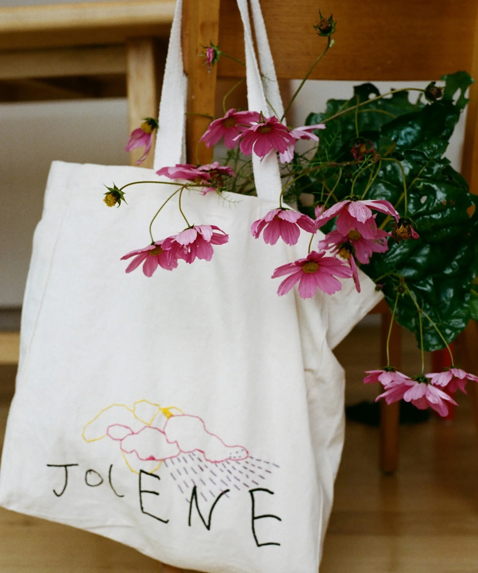 Off-white canvas tote bag featuring a stylized cloud and floral design, with pink cosmos flowers spilling out, resting on a wooden chair.
