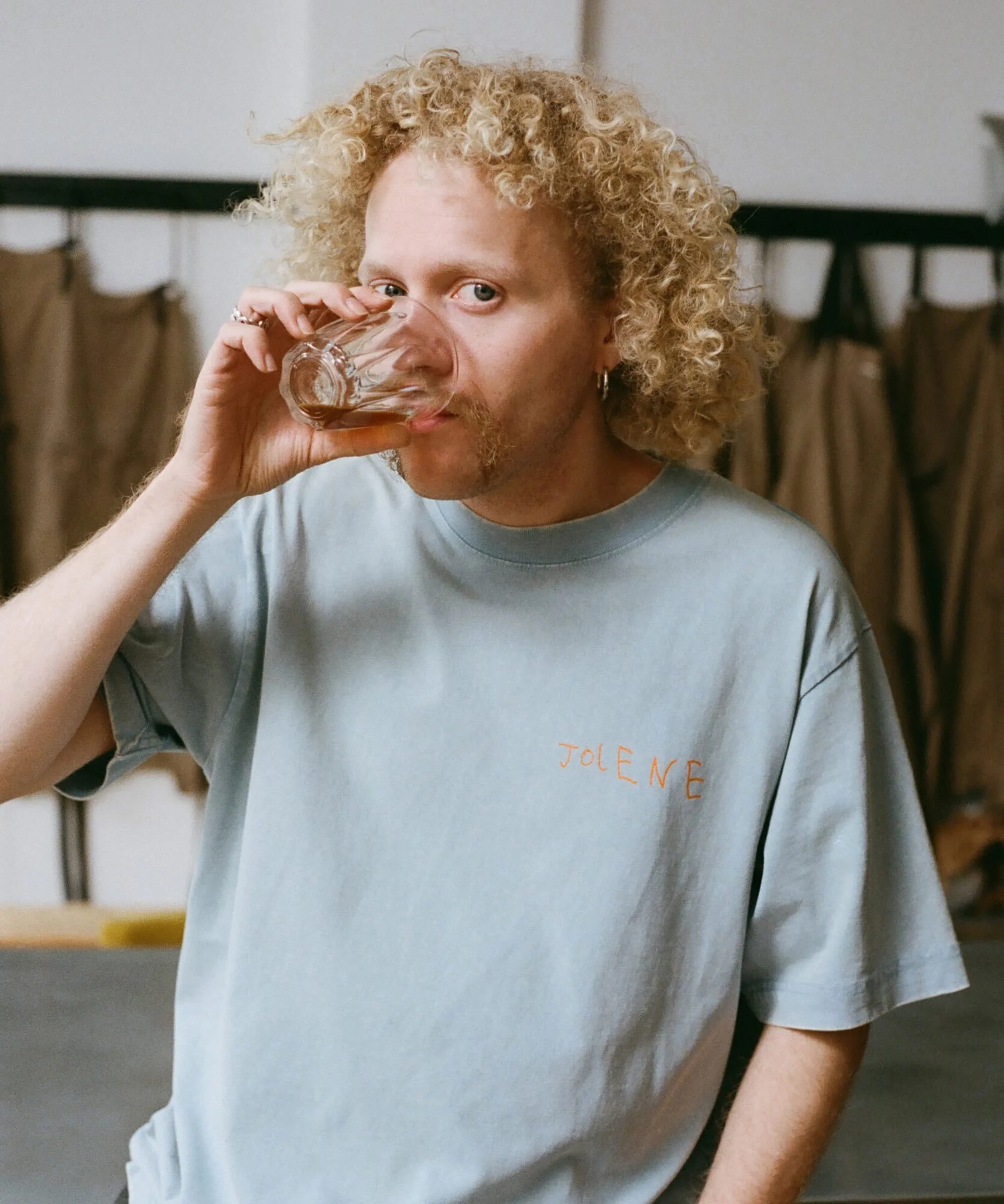 Adult male drinks from a clear glass in an indoor setting wearing a blue t-shirt with blonde curly hair and a mustache.