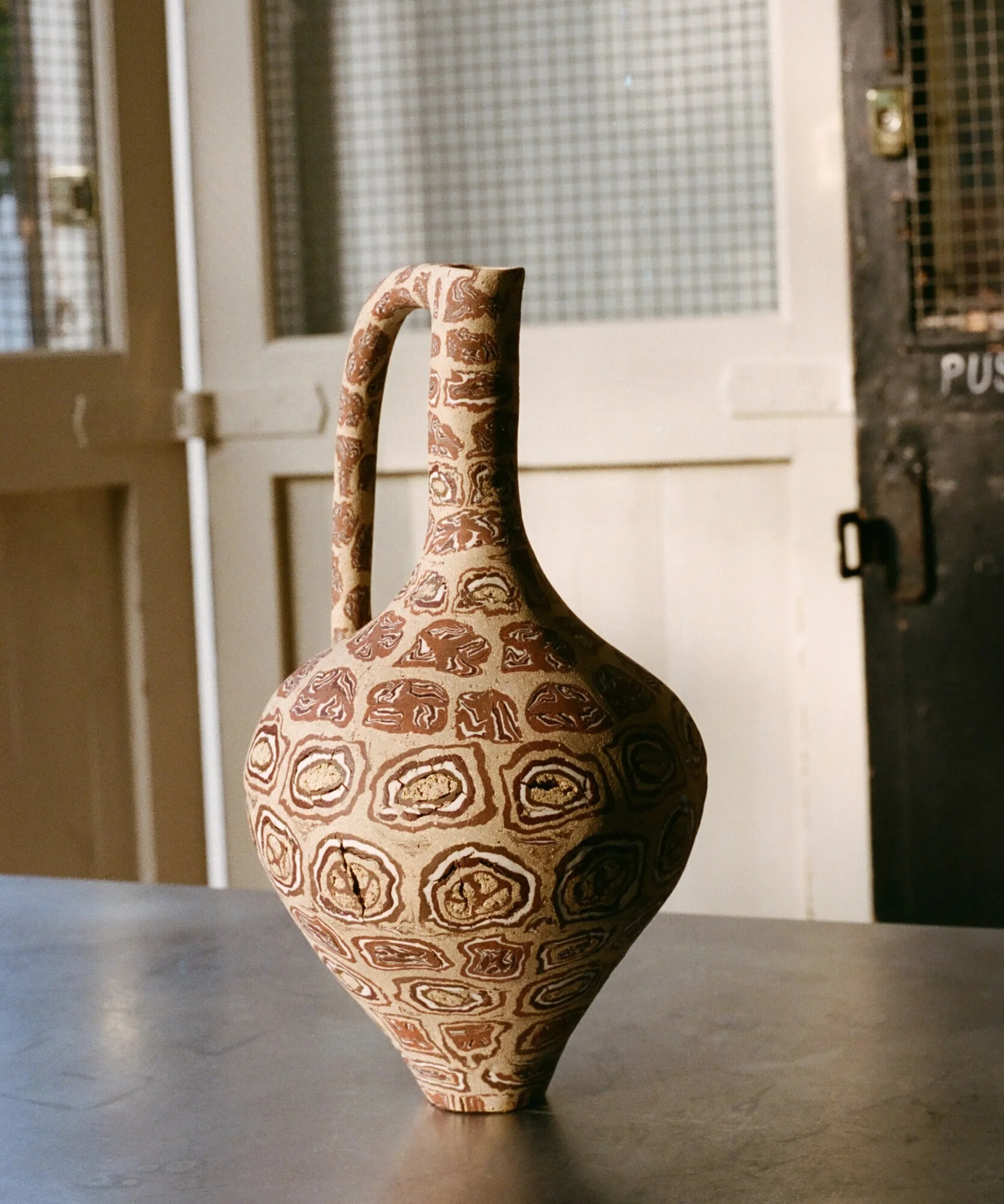 Ornate brown and beige patterned ceramic jug with handle stands on a table in front of doors with wired glass windows.