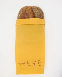 Loaf of seed-crusted bread in a yellow paper bag with handwritten text against a white background