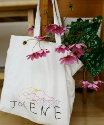 White canvas tote bag filled with pink cosmos flowers and green leaves hanging from a wooden chair.
