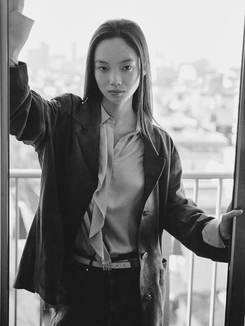 Elegant portrait of an Asian woman in a blazer standing near a window with a city view, showcasing fashion and urban lifestyle in black and white.