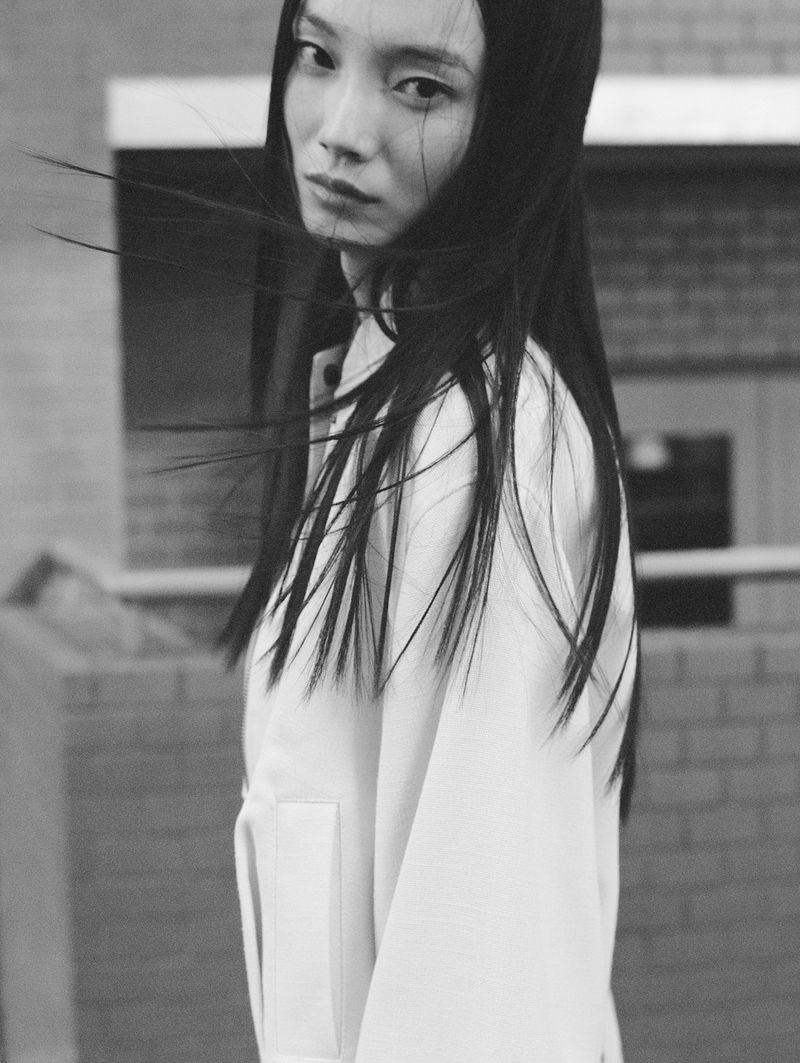 Monochrome portrait of an Asian woman with long dark hair blowing across her face wearing a white jacket outdoors against a brick wall