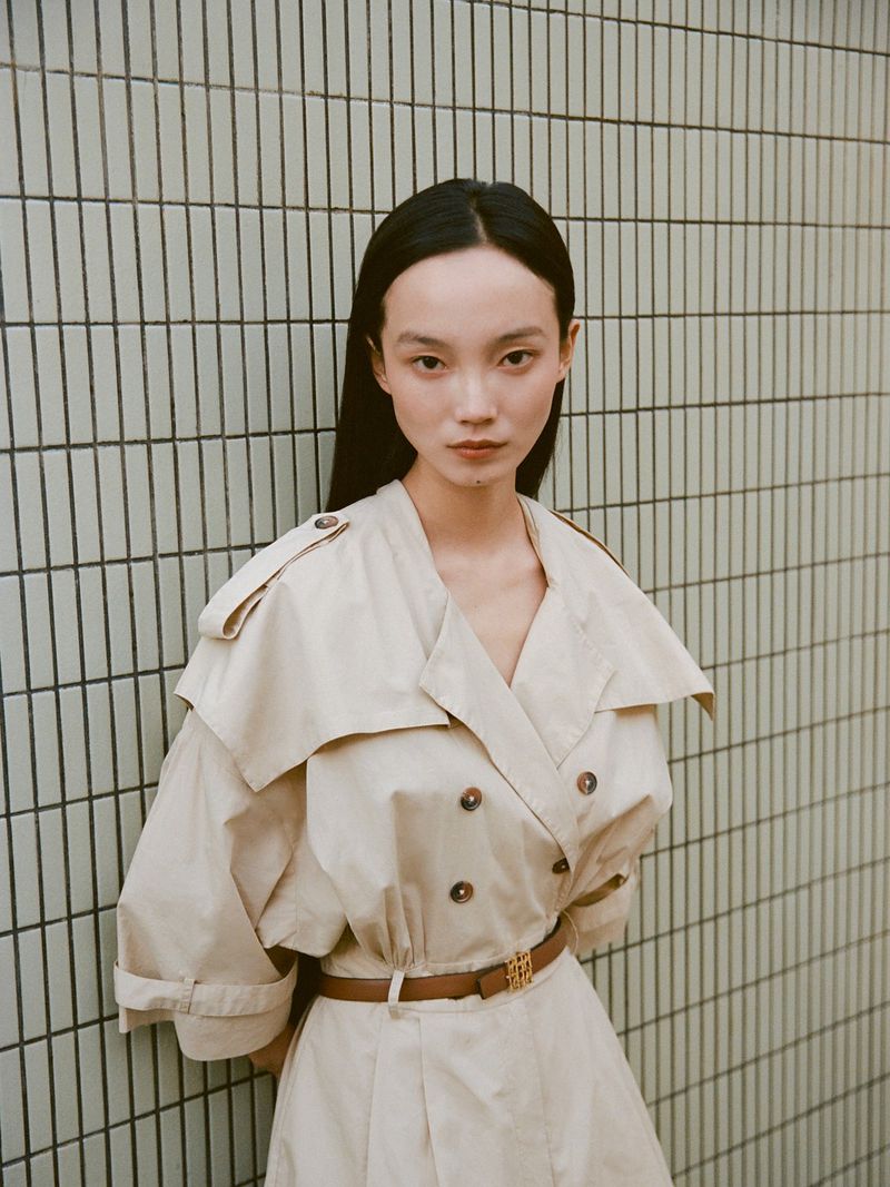 Asian woman wearing a cream belted trench coat posing against a tiled wall