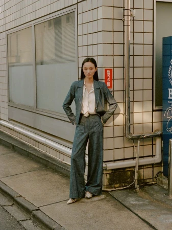 Woman in a denim suit stands on a city street in front of a building with white tiling and a window, next to a blue sign.