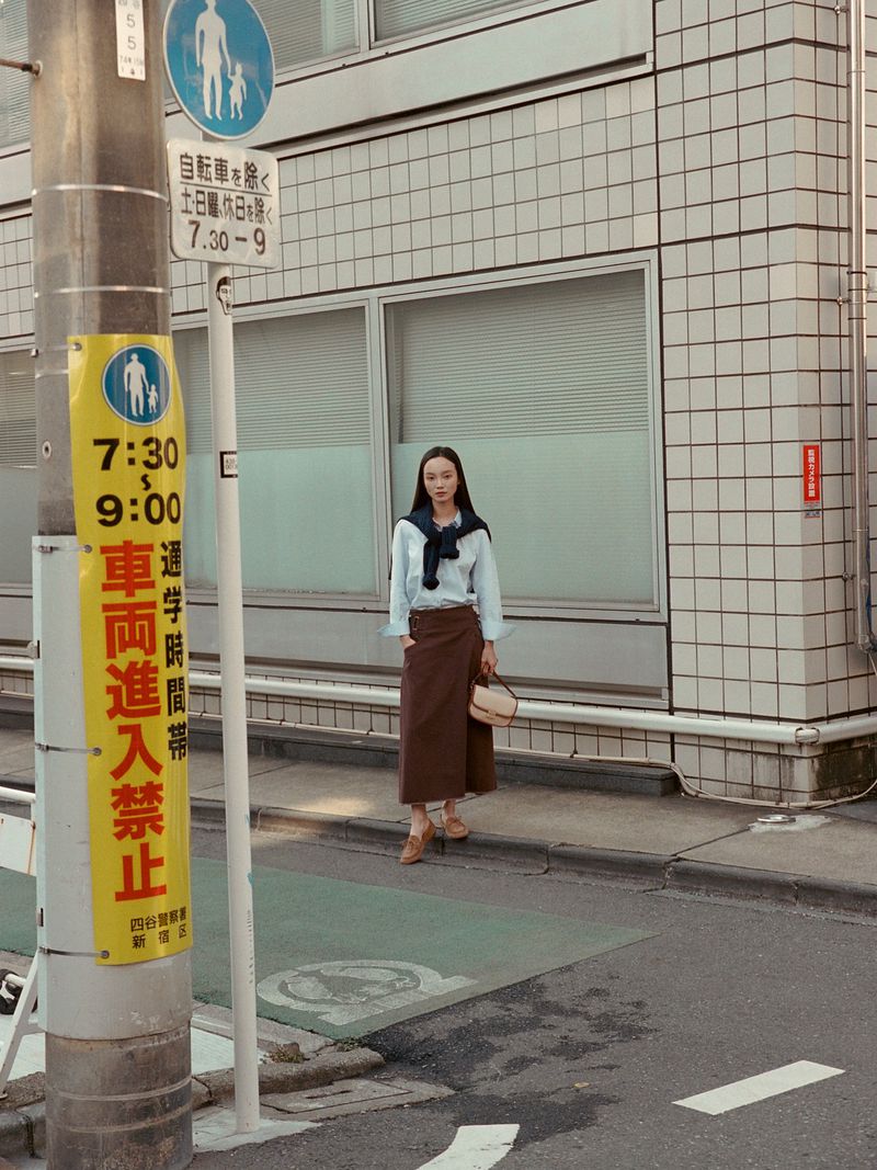 Adult woman in casual street style stands on sidewalk in front of building with pedestrian crossing sign and Japanese text.