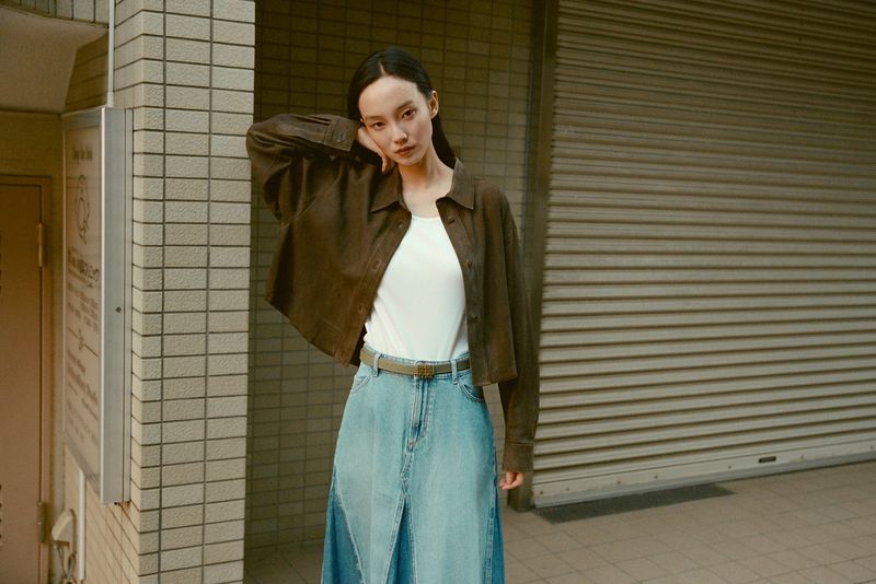An Asian woman poses in front of a neutral-colored building wearing a brown shirt jacket, white top, and long denim skirt with a belt.